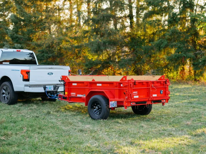 Red Wagon Rentals dump trailer loaded with lumber, hitched to a white Ford pickup at the edge of an autumn forest