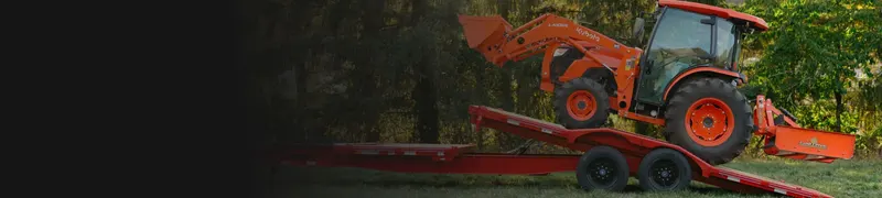 Orange tractor driving up the tilted deck of a red equipment trailer