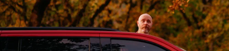 Andrew peeking over the roof of a red SUV in front of autumn trees